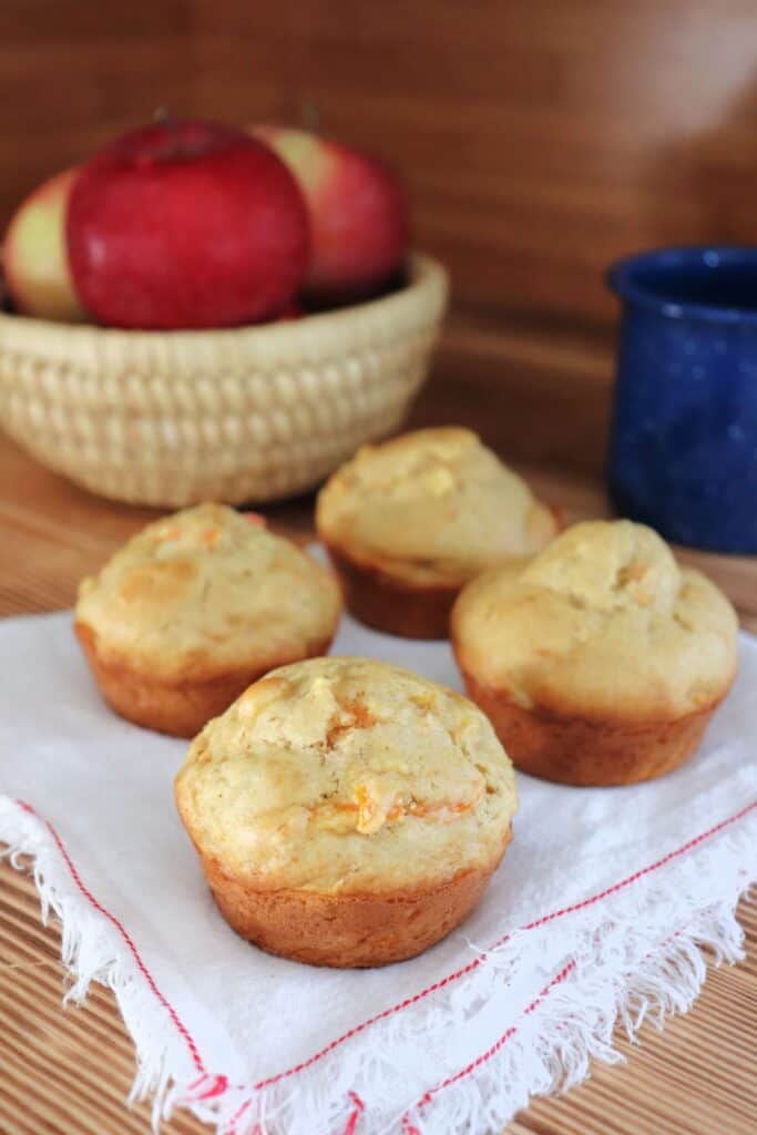 A close up of a muffin with more muffins behind it sitting on a white napkin. In the background is a blue coffee cup and a basket of fresh apples. 