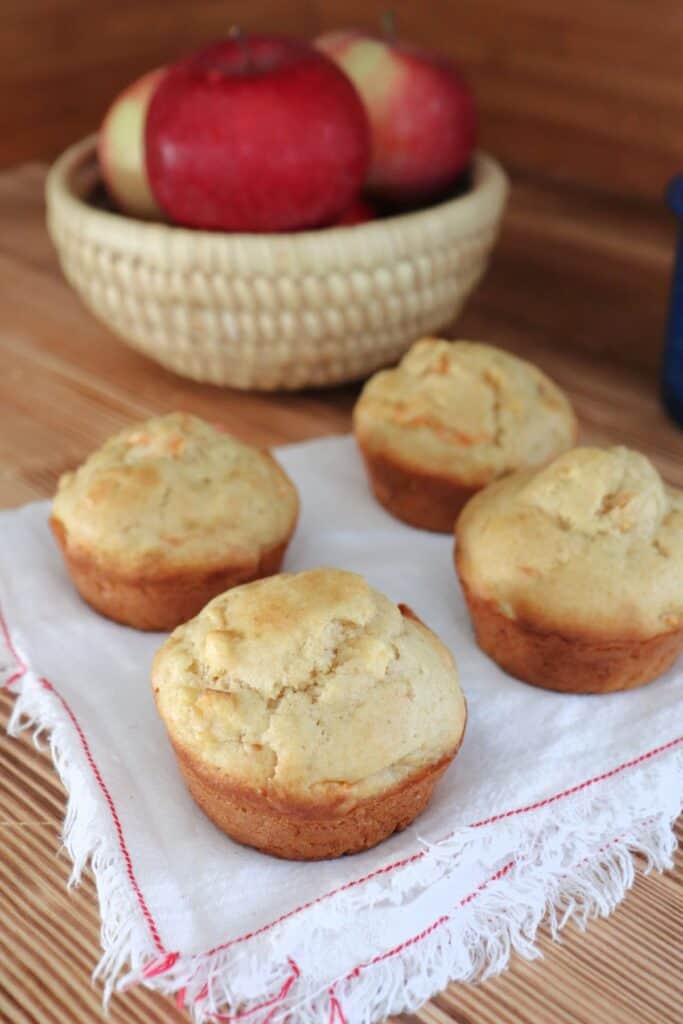 4 carrot apple muffins sitting on a white napkin. In the background is a basket of fresh red apples.