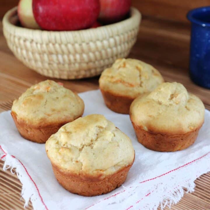4 muffins sitting on a white napkin with a basket of fresh apples in the background.