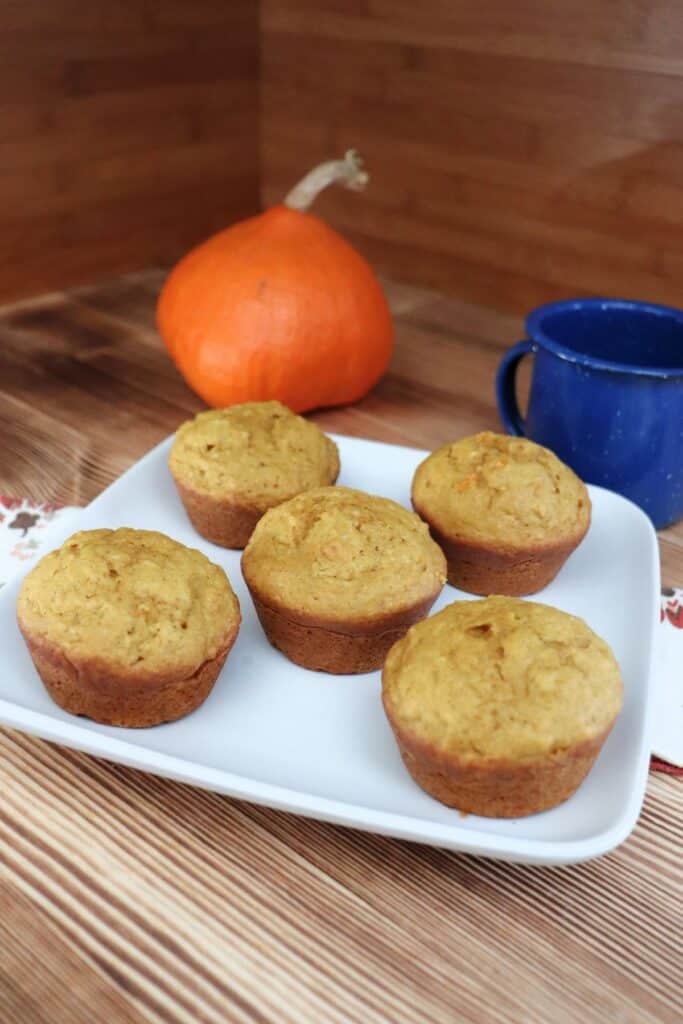A plate of muffins sits on top of a napkin. In the background a fresh pumpkin and blue tin cup. 