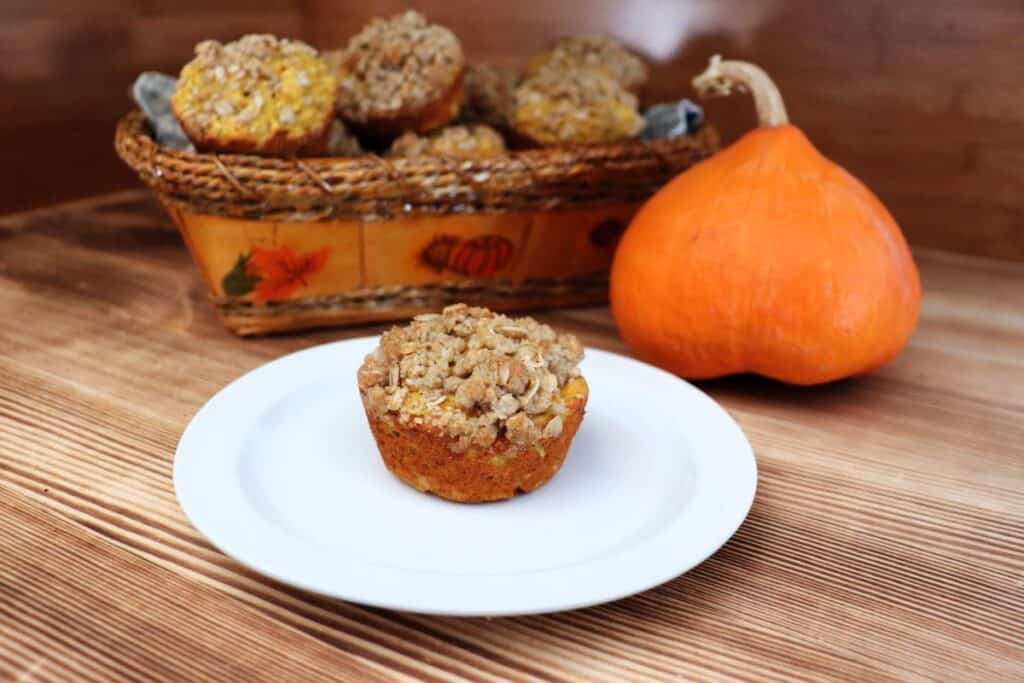 A muffin sits on a plate with a basket of more muffins and a pumpkin in the background. 