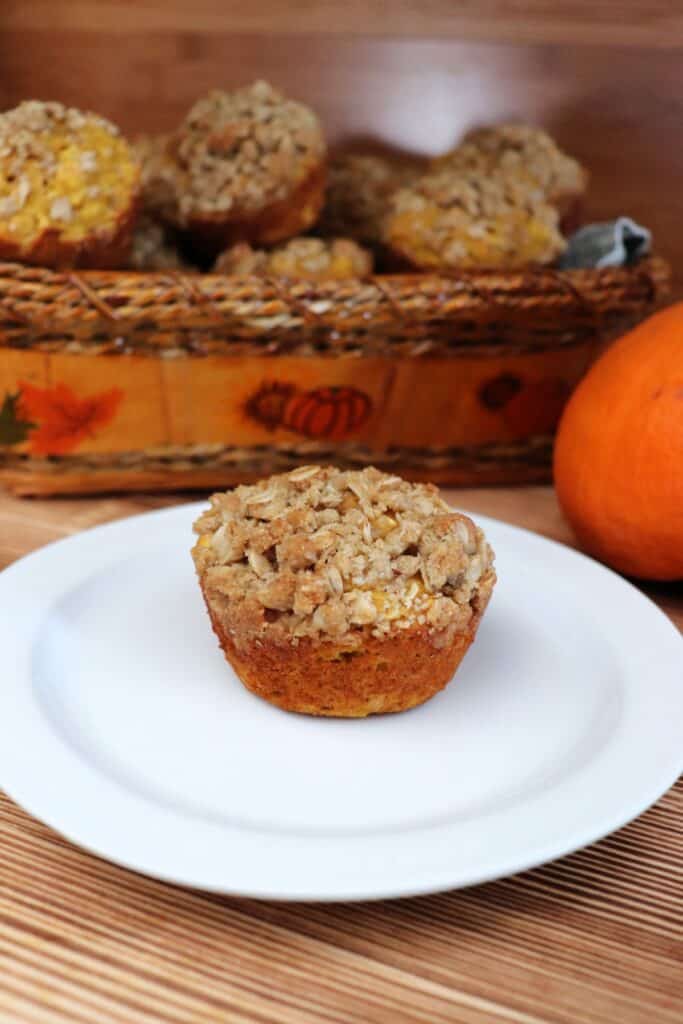 A muffin sits on a plate with a basket of more muffins and a pumpkin in the background. 