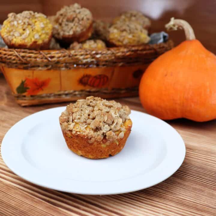 A muffin sits on a plate with a pumpkin and basket full of more muffins in the background.