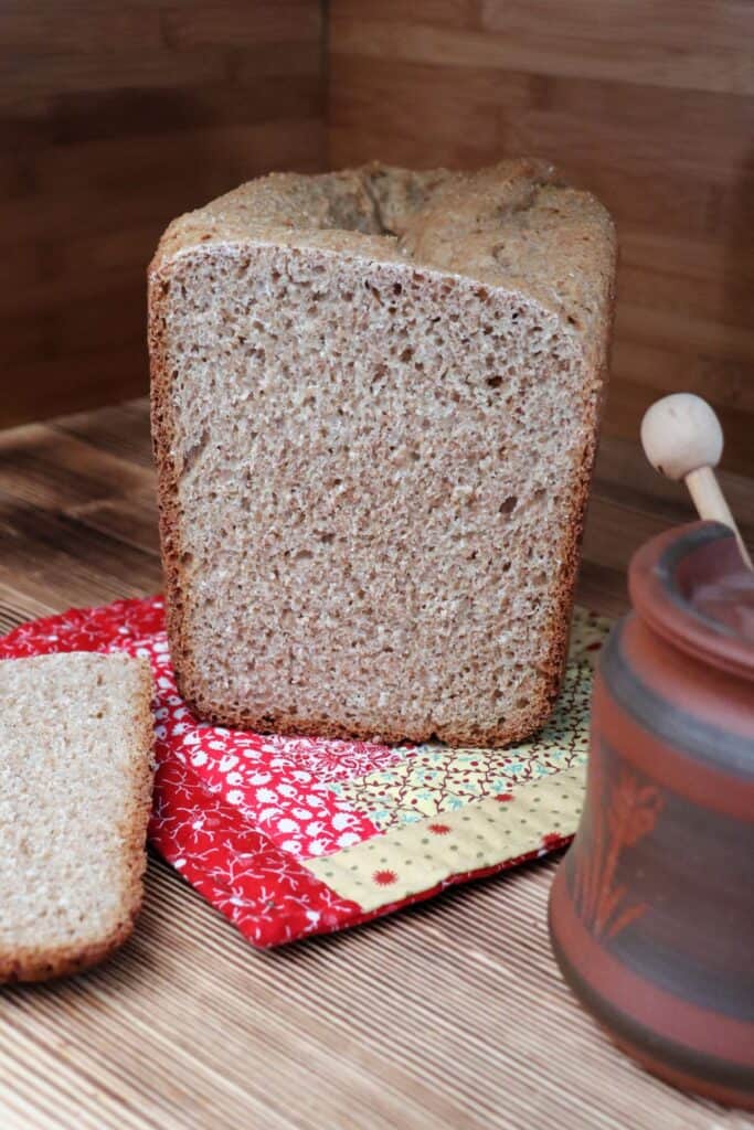 A loaf of bread with the end cuts off sits on a colorful cloth. In front of it is a slice of bread and a honey pot. 