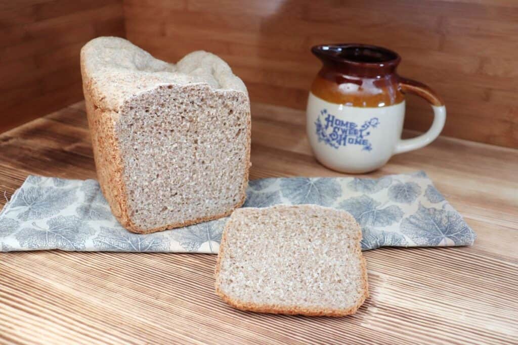 A slice of bread sits on a board with the remaining loaf sitting on a cloth behind it. An earthenware pitcher sits in the background. 