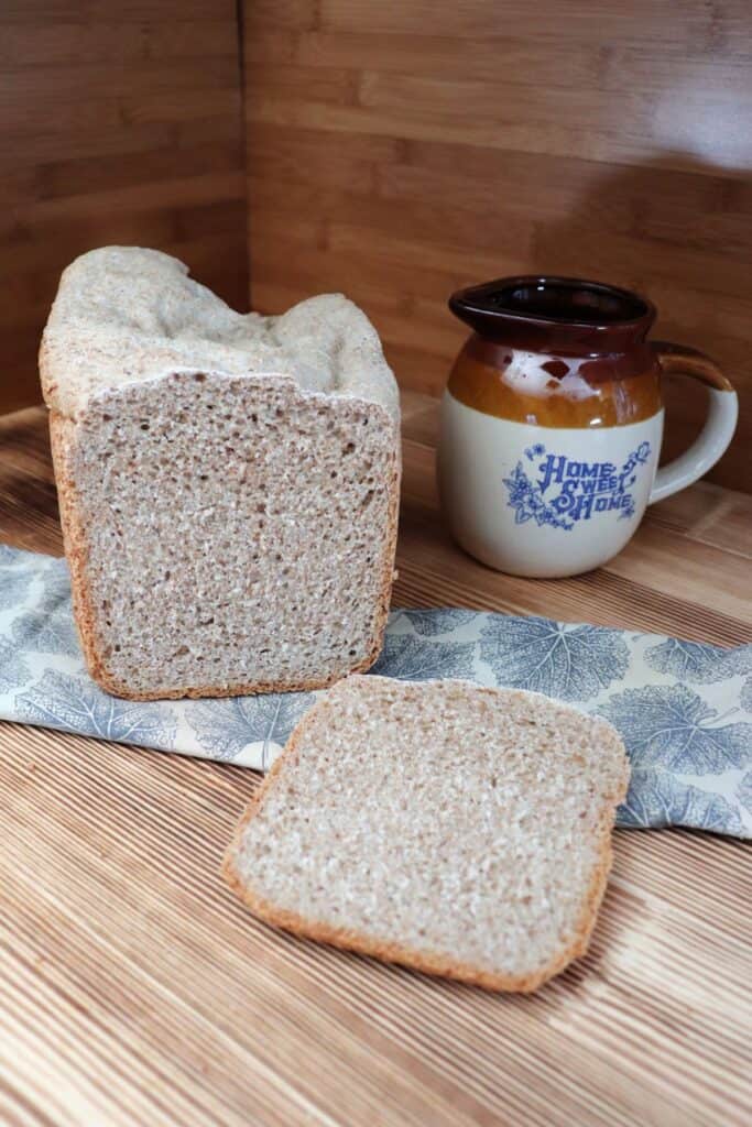 A slice of bread sits on a board with the remaining loaf sitting on a cloth behind it. An earthenware pitcher sits in the background. 