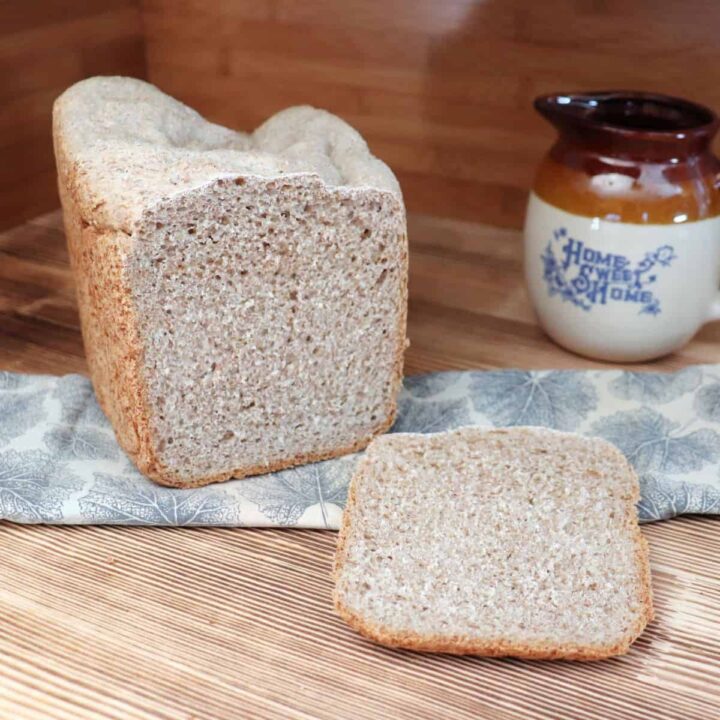 A slice of bread sits in front of the remaining loaf which is sitting on a napkin. An earthenware pitcher in the background.