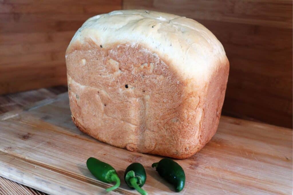 A loaf of bread sits on a cutting board with some fresh jalapeno peppers. 