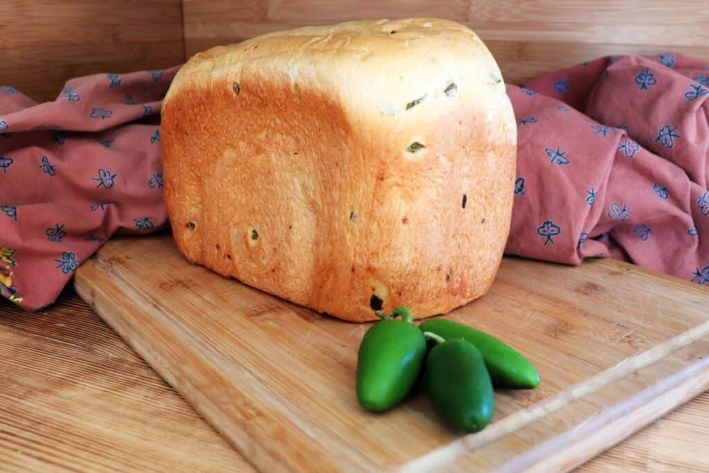 A loaf of cheddar jalapeno bread sits on a board with a red cloth behind it. Fresh jalapenos sit in front of the loaf. 