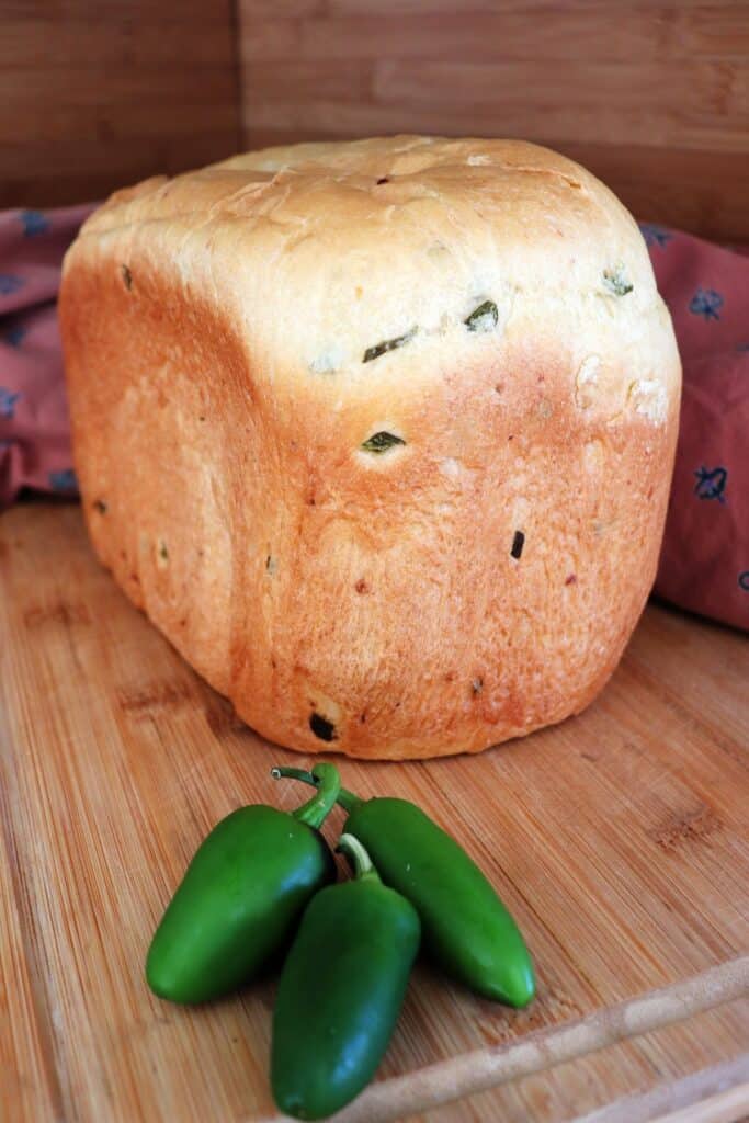 A loaf of cheddar jalapeno bread sits on a board with a red cloth behind it. Fresh jalapenos sit in front of the loaf. 