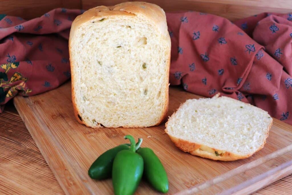 A loaf of bread with end cut off exposing pieces of green jalapeno peppers inside sits on a board. The heel sits next to it, a red cloth is in the background and fresh green jalapenos sit in front of the loaf. 