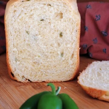 A loaf of bread with end cut off exposing pieces of green jalapeno peppers inside sits on a board. The heel sits next to it, a red cloth is in the background and fresh green jalapenos sit in front of the loaf.