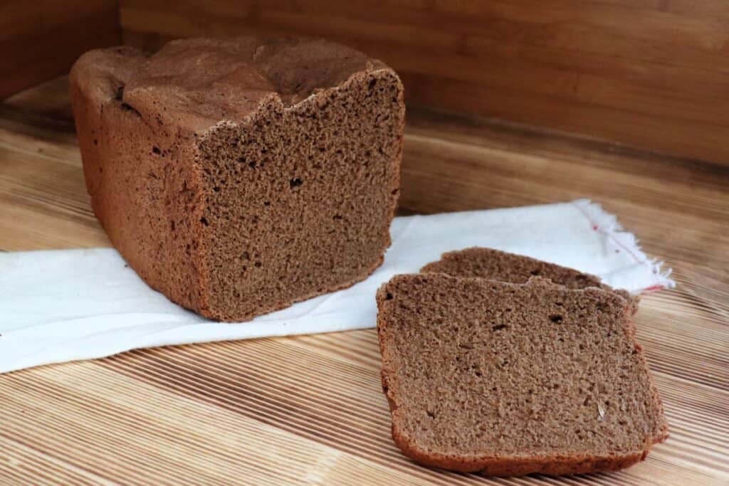 Slices of dark rye bread sit on a board with the remaining loaf sitting ona white cloth behind them. 