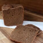 Slices of dark rye bread sit on a board with the remaining loaf sitting ona white cloth behind them.