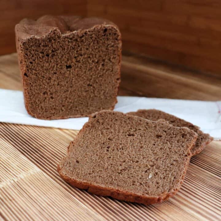 Slices of dark rye bread sit on a board with the remaining loaf sitting ona white cloth behind them.
