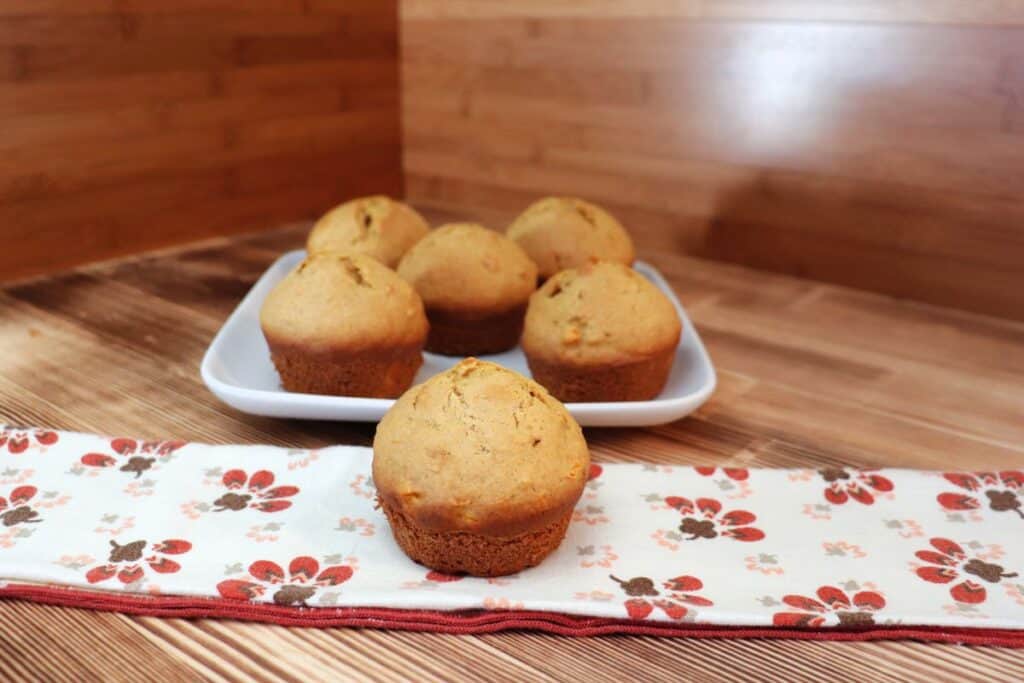 A muffin sits on a printed napkin with a platter of more muffins behind it.