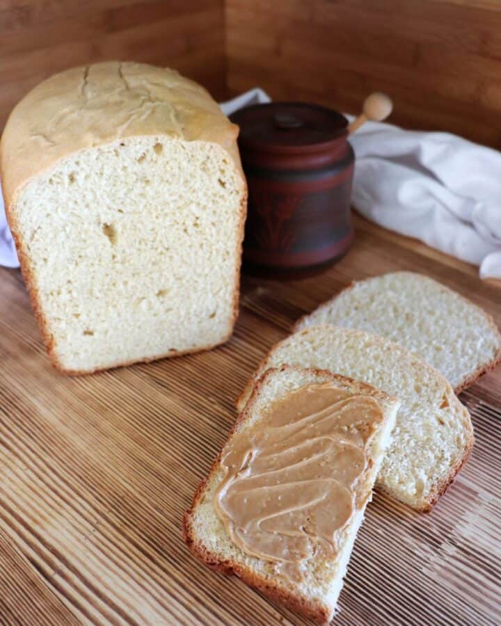 A slice of bread is slathered in peanut butter sitting on a board. The remaining loaf of white bread and a honey pot sit in the background.