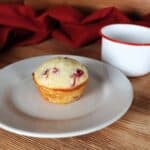 A cranberry muffin sits on a plate. A red and white coffee cup next to it. A red cloth is draped in the background.