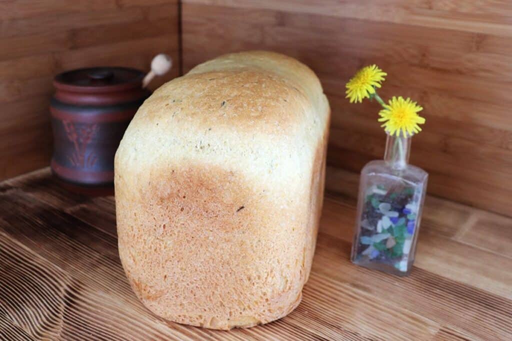 A loaf of bread sits on a board. To the left of the bread is a brown honey pot with the end of a dipper sticking out. To the right of the bread is a small bottle containing dandelion flowers.