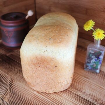 A loaf of bread sits on a board. To the left of the bread is a brown honey pot with the end of a dipper sticking out. To the right of the bread is a small bottle containing dandelion flowers.