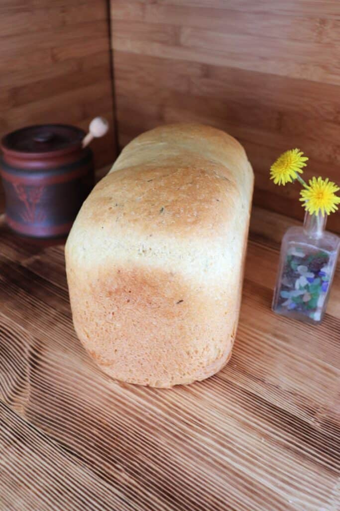 A loaf of bread sits on a board. To the left of the bread is a brown honey pot with the end of a dipper sticking out. To the right of the bread is a small bottle containing dandelion flowers.