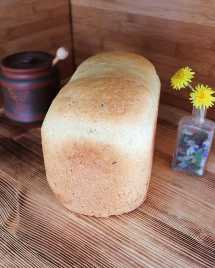 A loaf of bread sits on a board. To the left of the bread is a brown honey pot with the end of a dipper sticking out. To the right of the bread is a small bottle containing dandelion flowers.