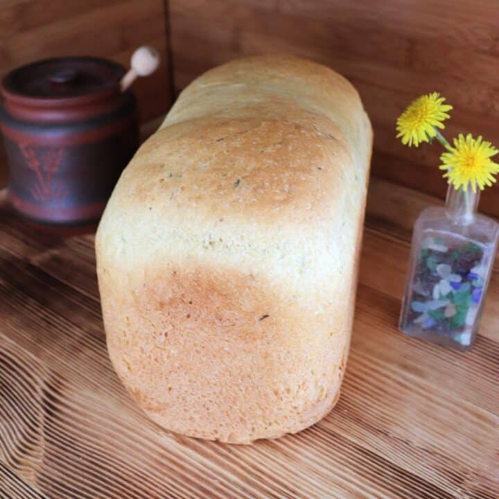 A loaf of bread sits on a board. To the left of the bread is a honey jar, to the right of the bread is a small bottle containing dandelion flowers.