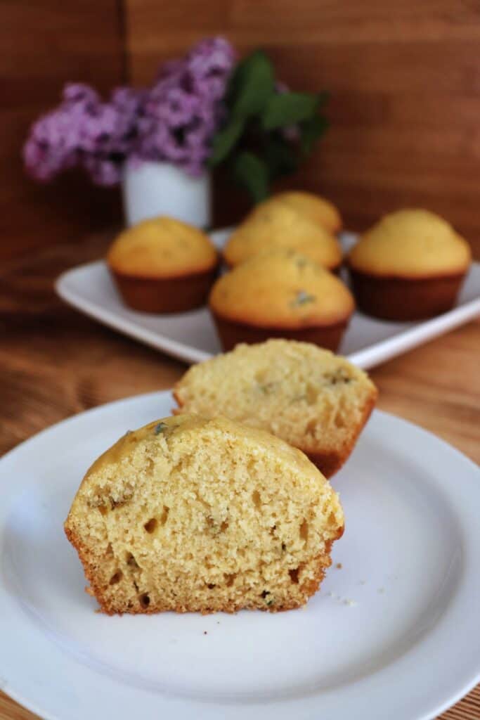 A muffin that has been in sliced in half sits on a white plate. In the background is a platter containing more muffins and a vase containing lilac flowers.