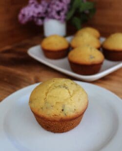 A muffin sits on a plate, bits of lilac flowers show inside the top of the baked muffin. In the background is a platter with more muffins and a white vase containing stems of fresh lilac flowers.