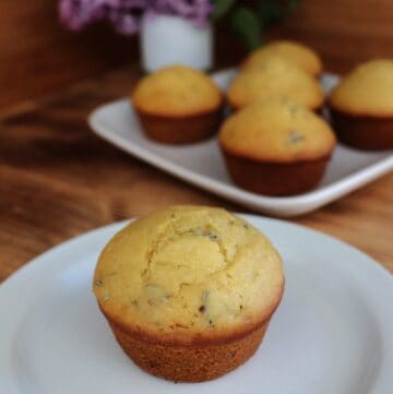A muffin sits on a plate, bits of lilac flowers show inside the top of the baked muffin. In the background is a platter with more muffins and a white vase containing stems of fresh lilac flowers.