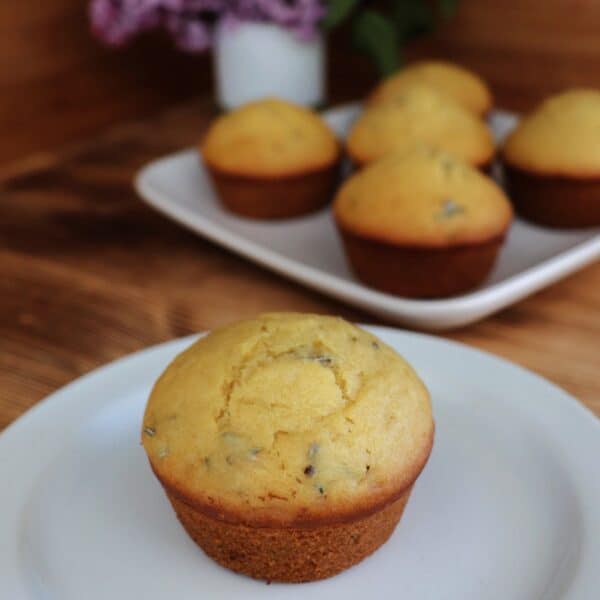 A muffin sits on a plate, bits of lilac flowers show inside the top of the baked muffin. In the background is a platter with more muffins and a white vase containing stems of fresh lilac flowers.