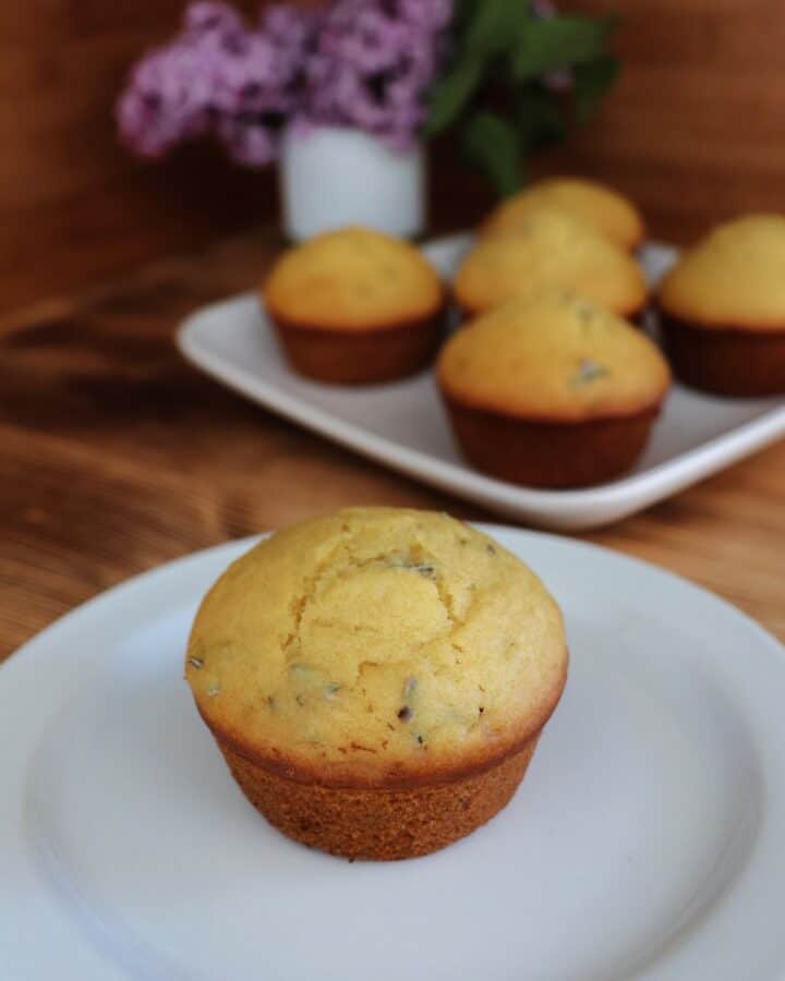 A muffin sits on a plate, bits of lilac flowers show inside the top of the baked muffin. In the background is a platter with more muffins and a white vase containing stems of fresh lilac flowers.
