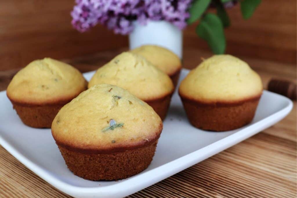 A platter of muffins sits on a table. The tops of the muffins show bits of flowers that were baked into them. In the background is a white vase full of purple lilac flowers.