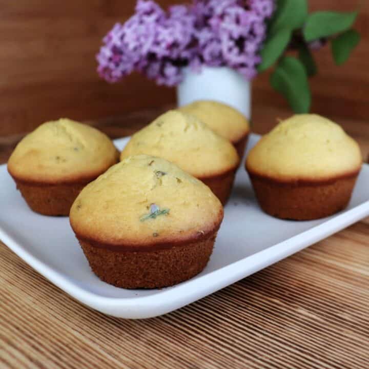 Muffins sit on a plate. The tops of the muffins show the lilac flowers cooked into them. In the background is a white vase full of fresh lilac flowers.