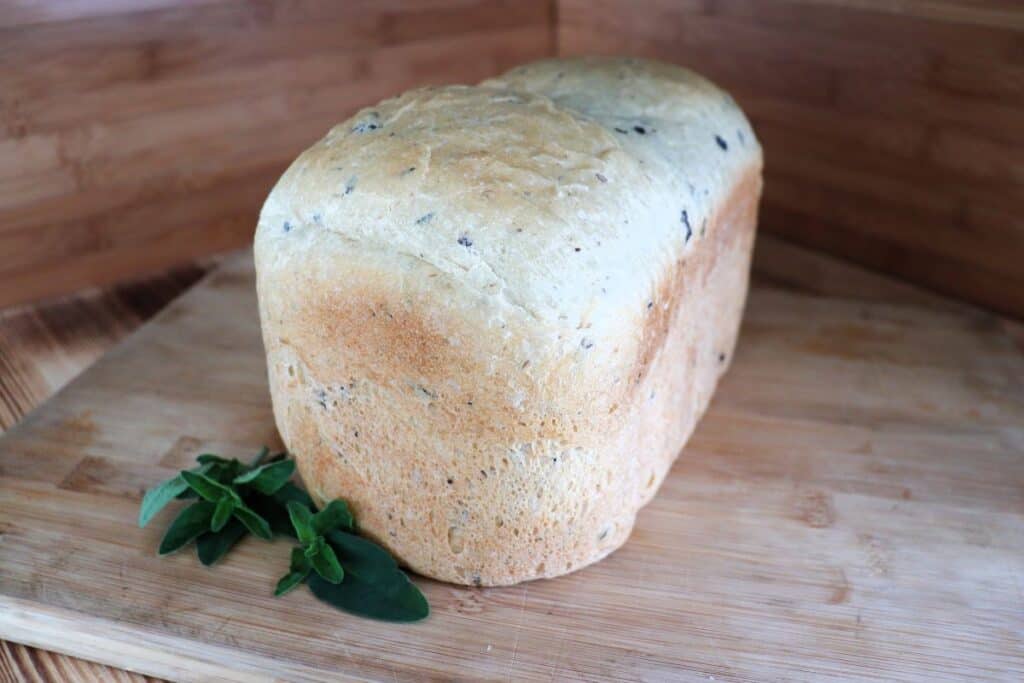 A loaf of bread studded with bits of black olives sits on a cutting board. A stem of fresh oregano sits next to it.