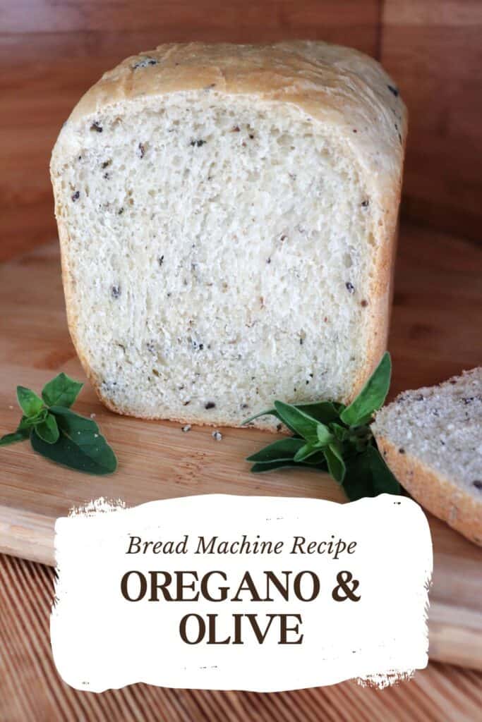 A loaf of bread with the end cut off exposing bits of black olives and chopped oregano inside sits on a cutting board. The cut off slice sits to the side and stems of fresh oregano surround the loaf. Text overlay reads: Bread Machine Recipe: Oregano & Olive.