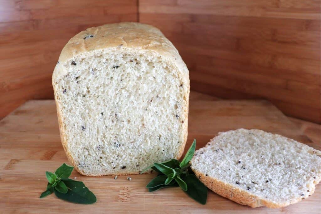A loaf of bread with the end cut off exposing bits of black olives and chopped oregano inside sits on a cutting board. The cut off slice sits to the side and stems of fresh oregano surround the loaf.