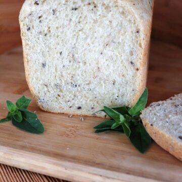 A loaf of bread with the end cut off exposing bits of black olives and chopped oregano inside sits on a cutting board. The cut off slice sits to the side and stems of fresh oregano surround the loaf.