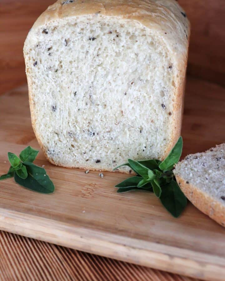 A loaf of bread with the end cut off exposing bits of black olives and chopped oregano inside sits on a cutting board. The cut off slice sits to the side and stems of fresh oregano surround the loaf.
