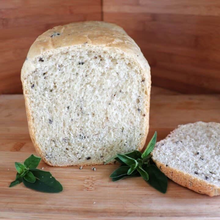A loaf of bread with the end cut off exposing bits of black olives and chopped oregano inside sits on a cutting board. The cut off slice sits to the side and stems of fresh oregano surround the loaf.