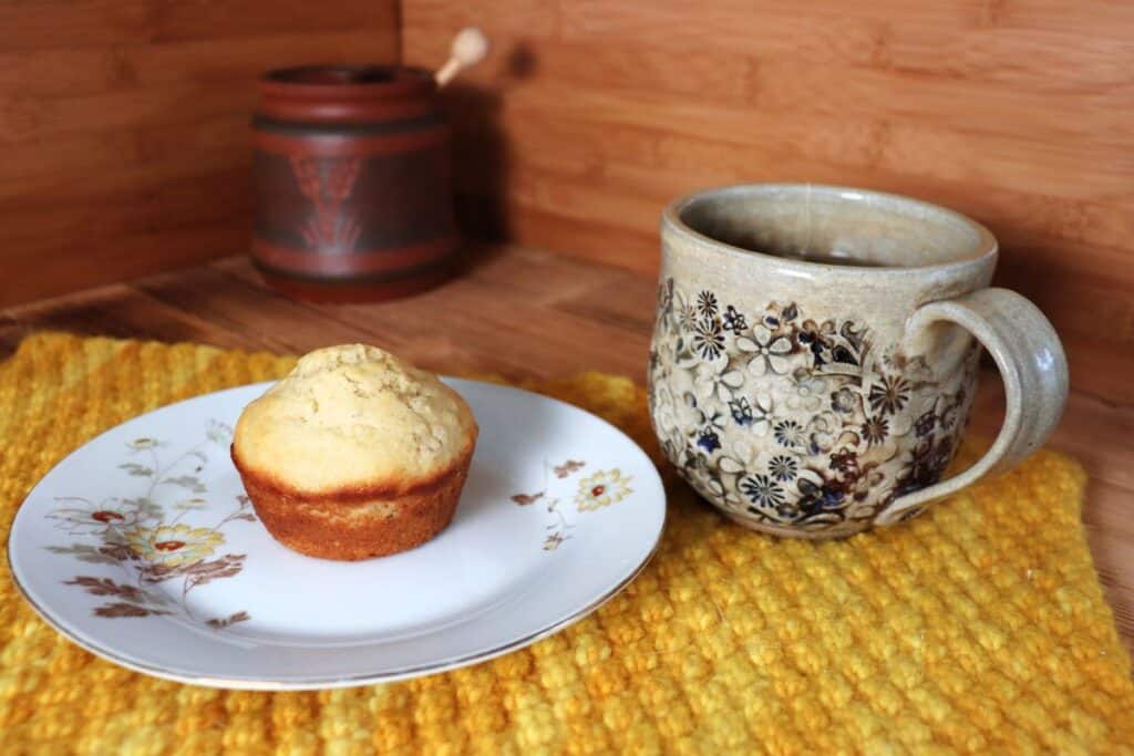 A muffin sit on a floral plate sitting on top of a yellow placemat.. A cup of tea sits next to it. In the background is a clay honey pot.
