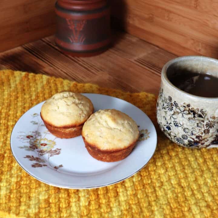 2 muffins sit on a floral plate sitting on top of a yellow placemat.. A cup of tea sits next to it. In the background is a clay honey pot.