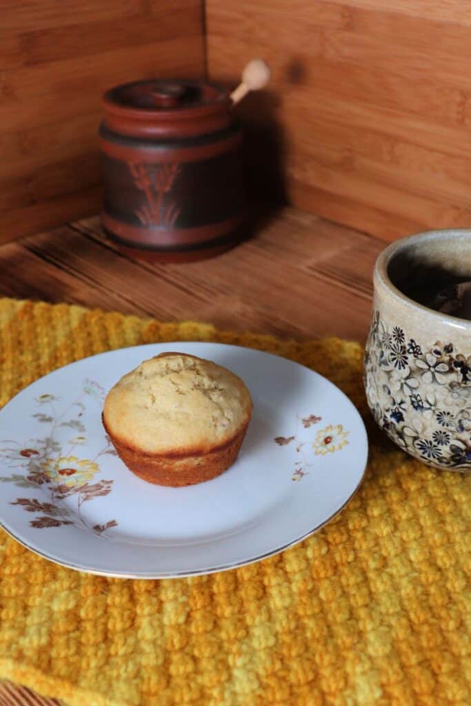 A muffin sit on a floral plate sitting on top of a yellow placemat.. A cup of tea sits next to it. In the background is a clay honey pot.