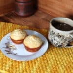2 muffins sit on a floral plate sitting on top of a yellow placemat.. A cup of tea sits next to it. In the background is a clay honey pot.
