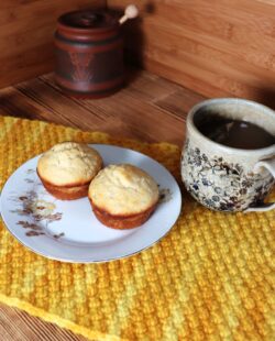 2 muffins sit on a floral plate sitting on top of a yellow placemat.. A cup of tea sits next to it. In the background is a clay honey pot.