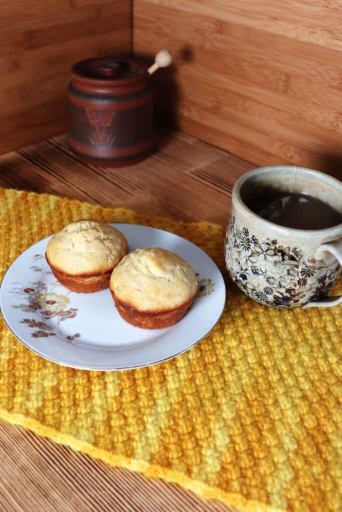 2 muffins sit on a floral plate sitting on top of a yellow placemat.. A cup of tea sits next to it. In the background is a clay honey pot.