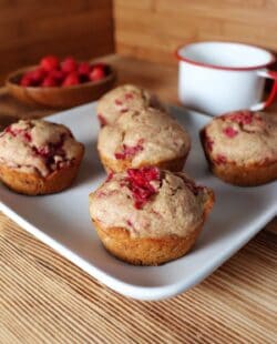 A square white plate with 5 raspberry spelt muffins sits on a counter. In the background is a white tin cup and a wooden bowl full of fresh raspberries.