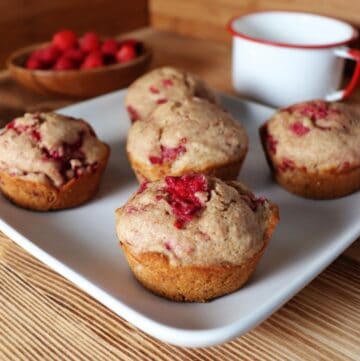 A square white plate with 5 raspberry spelt muffins sits on a counter. In the background is a white tin cup and a wooden bowl full of fresh raspberries.