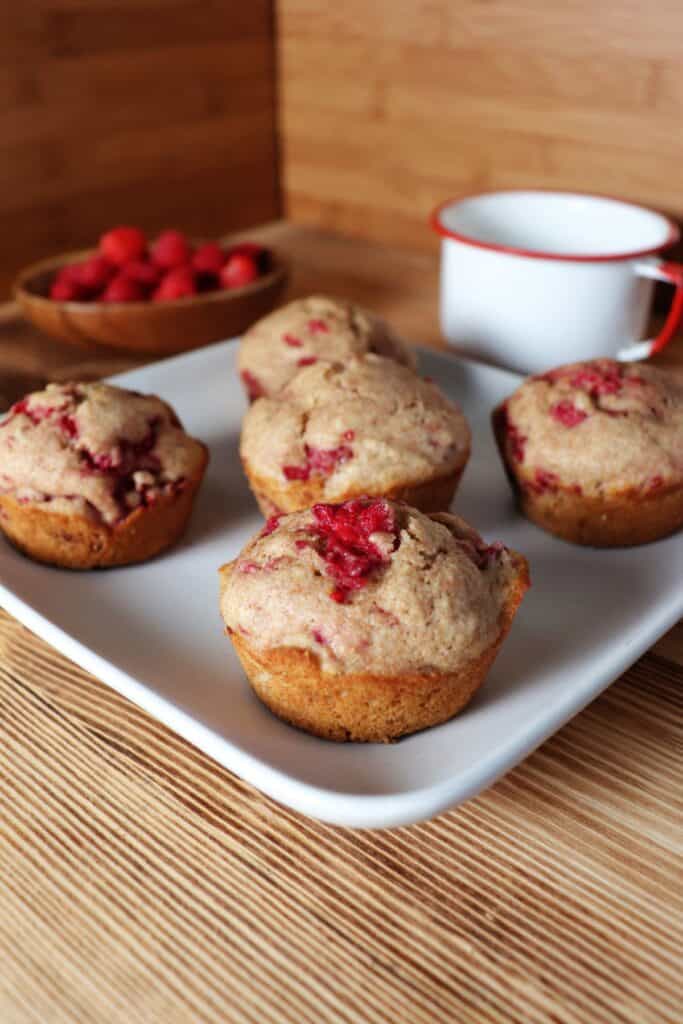 A square white plate with 5 raspberry spelt muffins sits on a counter. In the background is a white tin cup and a wooden bowl full of fresh raspberries. 