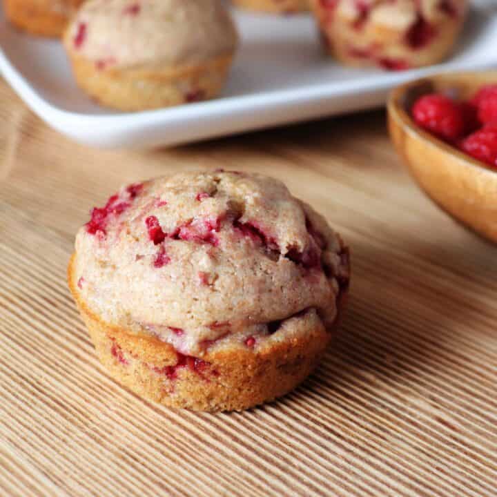 A raspberry spelt muffin sits on a board. In the background is small wooden bowl with fresh raspberries in it and a platter with more muffins.